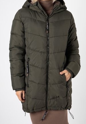 Persona che indossa una giacca a vento verde scuro fino al ginocchio con cappuccio, cerniera nera e mani in tasca sopra un vestito marrone a coste.