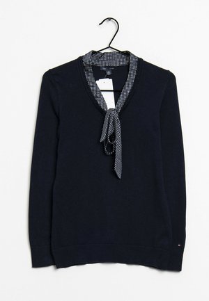 Pull bleu marine à manches longues avec un foulard à pois noué au niveau de l'encolure, suspendu sur un cintre noir sur fond blanc.