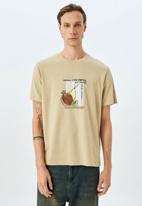Beige bomulds t-shirt med rund halsudskæring og korte ærmer. Har en farverig frugtgrafik på forsiden, herunder et granatæble og en avocado.