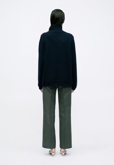 Navy turtleneck sweater met een gestructureerde afwerking, gecombineerd met groene geruite wijde broek. Model draagt beige puntige hakken.