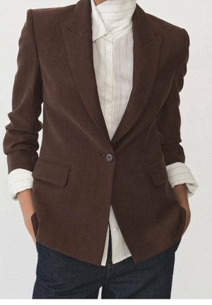 Personne portant un blazer marron à un seul bouton sur une chemise blanche à col avec manches retroussées et jean bleu foncé, mains dans les poches.