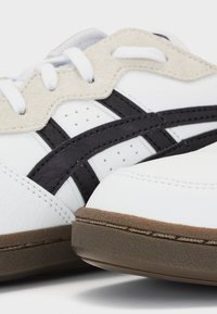Baskets blanches avec une tige en cuir texturé, des accents en daim beige, des bandes noires et une semelle en caoutchouc avec un bord marron.
