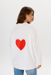 Weißes Langarmshirt mit einem großen roten Herz-Graphic auf der Rückseite, darüber der Text "KONZENTRIERE DICH AUF DEIN GLÜCK".