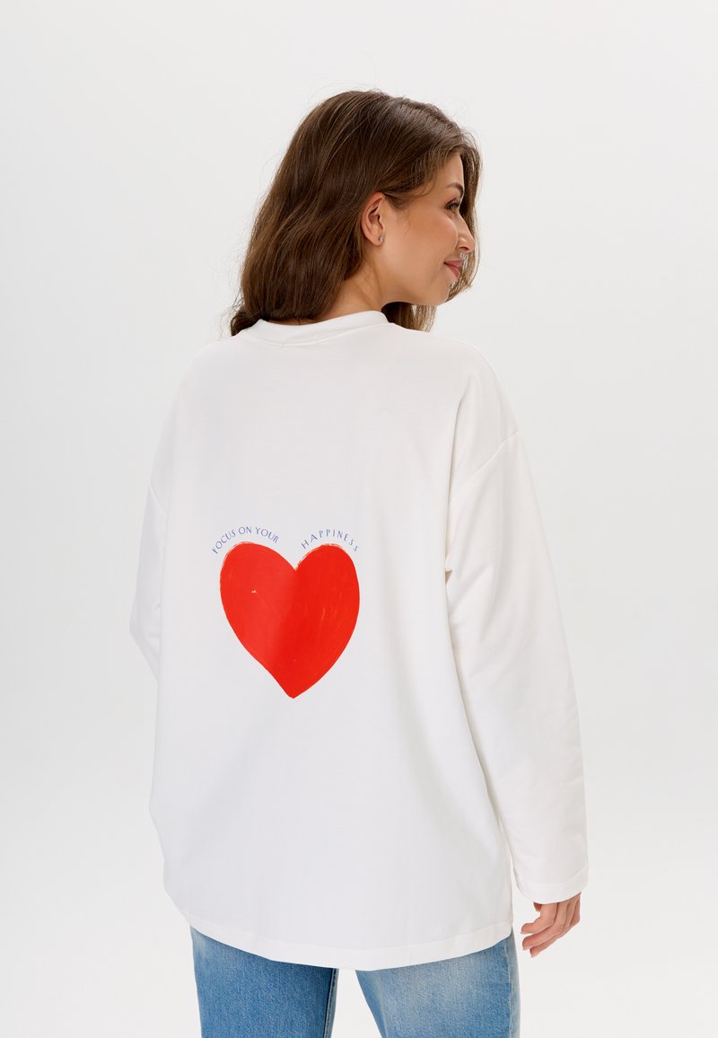 Weißes Langarmshirt mit einem großen roten Herz-Graphic auf der Rückseite, darüber der Text "KONZENTRIERE DICH AUF DEIN GLÜCK".
