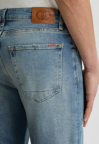 A világoskék farmernadrág övén egy bőr márkás folt található, és két hátsó zseb van, enyhén kopott részletekkel.