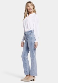 Blusa blanca de manga larga con pliegues, combinada con jeans de mezclilla acampanados en azul claro. La modelo lleva zapatos de tacón cuadrado beige.