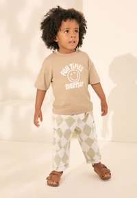 Lyhythihainen T-paita, jossa valkoisella teksti "FUN TIMES EVERYDAY", yhdistettynä löysiin valkoisiin housuihin, joissa on vihreitä timanttikuvioita, ja ruskeisiin sandaaleihin.