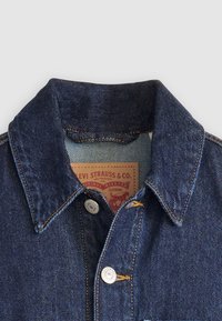 Mørk denimjakke krage med sølv snapknapper, kontrasterende gul søm, og en brun label med merkeinformasjon inni.