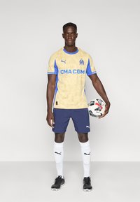 Κίτρινη και μπλε ποδοσφαιρική φανέλα με κοντά μανίκια, με λεπτομερή σχέδια, σε συνδυασμό με ναυτικό σορτς και μαύρα ποδοσφαιρικά παπούτσια, κρατώντας μια μπάλα ποδοσφαίρου.