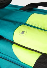Türkise Rucksack mit einem leuchtend gelben Mittelteil, strukturiertem Stoff, schwarzen Reißverschlüssen und einem kreisförmigen Logodetail auf dem gelben Bereich.