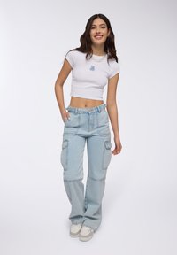 T-shirt bianca corta con logo di orso blu, abbinata a pantaloni cargo larghi azzurri con multiple tasche e una vita alta.