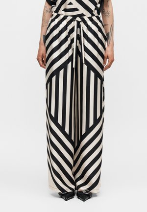 Spodnie o szerokich nogawkach z odważnymi czarno-białymi geometrycznymi paskami, elastyczny pas ze sznurkiem, noszone z czarnymi spiczastymi butami i widocznymi tatuażami na ramionach.