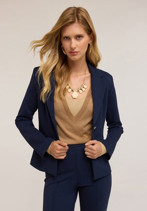 Blazer blu navy con una vestibilità su misura, abbinato a un top in maglia metallica dorata e una collana in oro massiccio. Texture morbide e linee pulite.