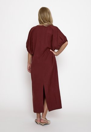 Donna con capelli biondi che indossa un abito midi bordeaux con maniche a sbuffo e sandali beige con tacco basso, in piedi di spalle con la mano sul fianco.