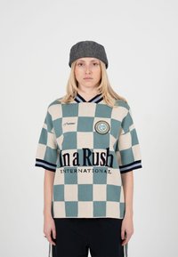 Ung person iført en ternet skjorte i teal og beige med teksten "In a Rush International" og en grå flat kasket, stående mod en ensfarvet baggrund.