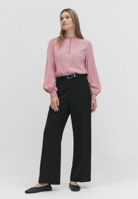 Vaaleanpunainen pitkähihainen paita napituksella ja rypytetyillä hihansuilla, yhdistettynä korkeavyötäröisiin mustiin leveälahkeisiin housuihin ja mustiin balettitossuihin.