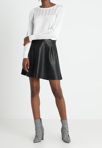 Pull en maille blanche avec texture à motifs, jupe évasée en similicuir noir, et bottines grises à carreaux avec talons blocs.