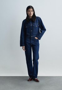 Kurtka dżinsowa w ciemnoniebieskim kolorze, z futrzanym kołnierzem i przednimi kieszeniami, zestawiona z dopasowanymi niebieskimi dżinsami i bordowymi butami na betonowej podłodze.