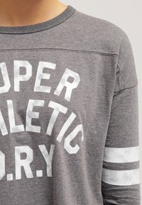 Grå långärmad tröja med vit text "SUPER ATHLETIC D.R.Y." och vita ränder på ärmarna. Mjuk tygkvalitet med rundad halsringning.