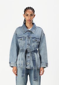 Jeansjakke med belte i midjen, oversized passform, to brystlommer og stor krage. Lys blå vask med en teksturert finish.