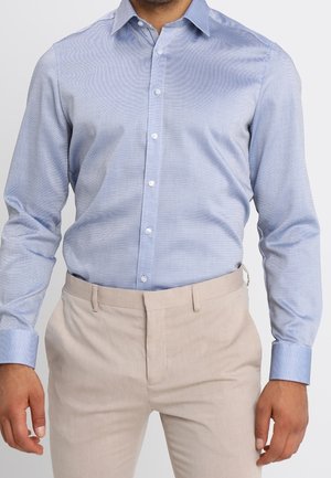 Mann trägt ein hellblau gemustertes Hemd mit langen Ärmeln, in beige Hosen gesteckt, die bis zur Mitte des Oberschenkels gekürzt sind.