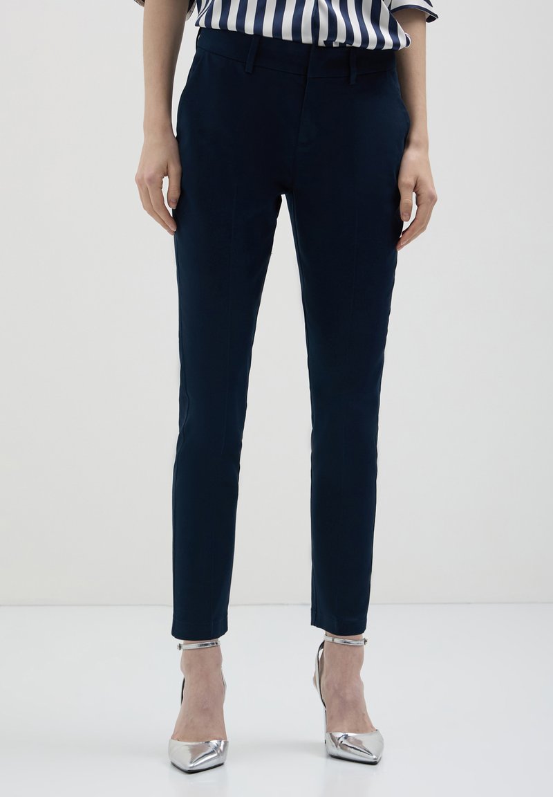 Pantaloni tailored blu navy con una vestibilità slim, caratterizzati da una superficie liscia, tasche frontali e passanti per cintura, abbinati a scarpe con tacco alto in argento.