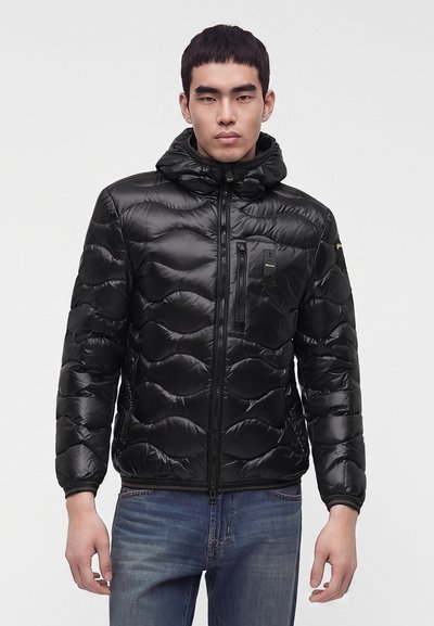 Jeune homme portant une veste matelassée noire brillante avec une capuche et une fermeture éclair à l'avant, associée à un jean bleu, se tenant devant un fond uni.