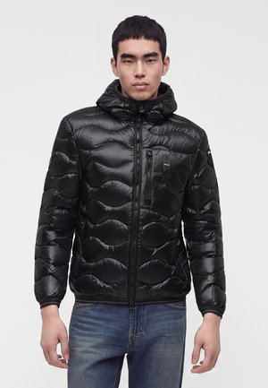 Jeune homme portant une veste matelassée noire brillante avec une capuche et une fermeture éclair à l'avant, associée à un jean bleu, se tenant devant un fond uni.