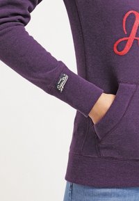 Sudadera morada con puños y dobladillo acanalados, que presenta una letra "J" roja bordada en el pecho y una etiqueta en la manga.
