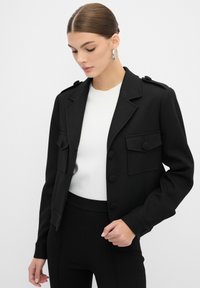 Czarna krótka kurtka o strukturalnym designie, z dwoma przednimi kieszeniami, zapięciem na guziki i epoletami na ramionach. Gładka tekstura.