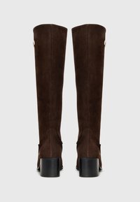 Stivali al ginocchio in suede marrone con tacco a blocco, caratterizzati da dettagli di cucitura curvi e un accento decorativo a forma di bottone vicino al polpaccio esterno.