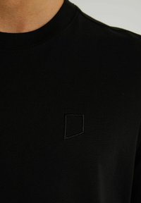 Tricou negru din bumbac neted, cu un guler rotund cu nervuri și un logo brodat subtil în negru în partea stângă jos.