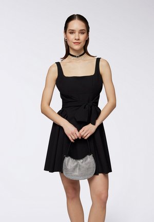 Abito nero senza maniche con scollatura quadrata, corpetto aderente e dettaglio a fiocco in vita. La modella tiene una borsa a tracolla metallic a argento con superficie in gioiello.