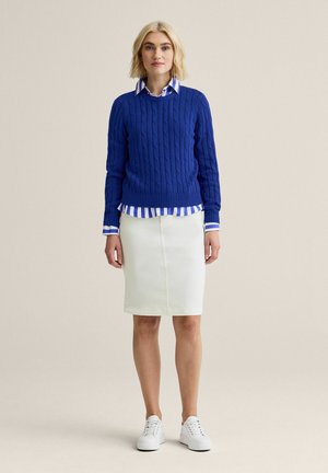 Kvinde står iført en blå kabelstrikket sweater over en blå- og hvidstribet skjorte, hvid knælang nederdel og hvide sneakers.
