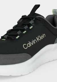 Μαύρο και γκρι παπούτσι Calvin Klein με λευκή σόλα, μαύρα κορδόνια και ανοιχτό πράσινες λεπτομέρειες στα παπούτσια και στη θηλιά της φτέρνας.