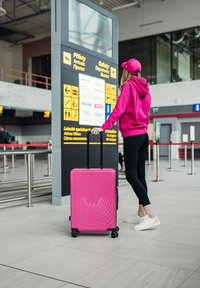 Světlý růžový kufr s tvrdou skořepinou a vlnkovým vzorem, vybavený teleskopickou rukojetí a čtyřmi koly, stojí vedle cestovatele.