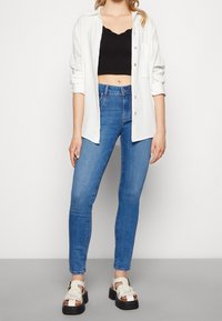 Kvinna som bär blå högmidjade skinny jeans, svart kort topp, vit skjorta i knappar och vita sandaler med svarta sulor.