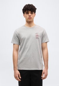 Νέος με κατσαρά μαλλιά που φοράει ανοιχτό γκρι μπλουζάκι με μικρό κοκκινωπό σχέδιο στο αριστερό στήθος και μαύρο παντελόνι.