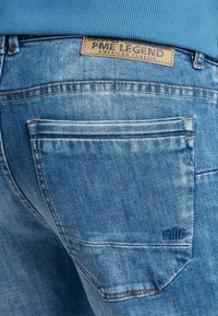 Modre kavbojke iz denima z teksturiranim zaključkom, ki vključujejo rjavo usnjeno oznako in kontrastno šivanje. Imajo podrobnosti zadnjega žepa in logotip.