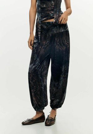 Osoba oblečená v čiernych vzorovaných voľných nohaviciach s elastickými manžetami, v kombinácii s tmavohnedými šnurovacími topánkami na plochom podpätku, stojaca na jednoduchom pozadí.