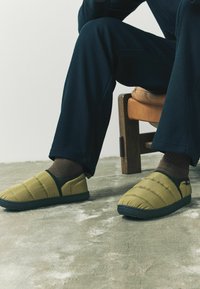 Kvalitné zelené croc topánky s čiernymi akcentmi, texturovaná tkanina a flexibilná podrážka, nosené s hnedými ponožkami a námorníckymi nohavicami, sedenie na stoličke.