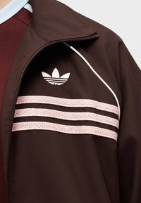 Κοντινό πλάνο ενός ατόμου που φοράει καφέ μπουφάν Adidas με τρεις ροζ υφασμάτινες ρίγες και λογότυπο Adidas trefoil σε λευκό στο στήθος.