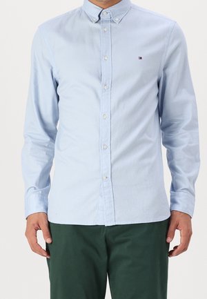 Mann trägt hellblaues, langärmeliges Hemd mit Knöpfen und kleinem Logo auf der Brust sowie dunkelgrüne Hose, steht vor einem einfarbigen Hintergrund.