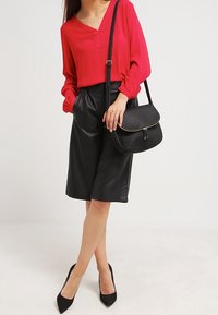Donna che indossa una blusa rossa, una gonna in pelle nera fino al ginocchio, tacchi neri e porta una borsa a tracolla nera.