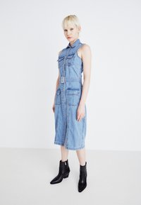 Abito di denim con vita cintata, design senza maniche, tasche frontali e chiusura a bottoni. Indossato con stivali neri alla caviglia con dettagli borchiati.