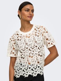 Femeie purtând un top alb cu dantelă florală cu mânecă scurtă peste o haină neagră, pozând cu o mână pe șold, fundal neutru.