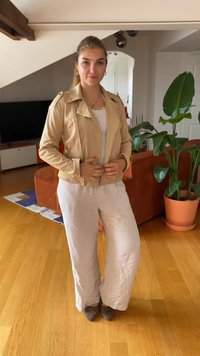 Beige Kunstlederjacke mit silbernen Reißverschlüssen über einem weißen Oberteil, kombiniert mit lockeren, hellgrauen Leinenhosen und grauen Schuhen. Innenaufenthalt.
