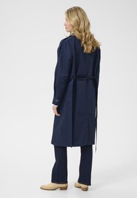 Navyblauwe trenchcoat van waterdicht materiaal, met een ceintuur in de taille, lange mouwen en schouderepauletten. Draagt over donkere flared jeans.