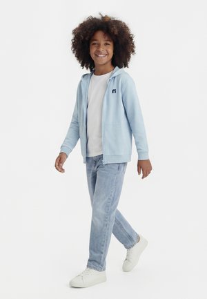 Smiling child walking, wearing a light blue zip-up hoodie, white t-shirt, light blue jeans, and white sneakers on a white background.