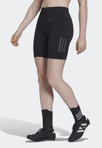 Shorts de sport noirs avec une taille haute, fabriqués en matériau extensible, avec trois bandes grises sur le côté. Portés avec des chaussettes et des chaussures noires.